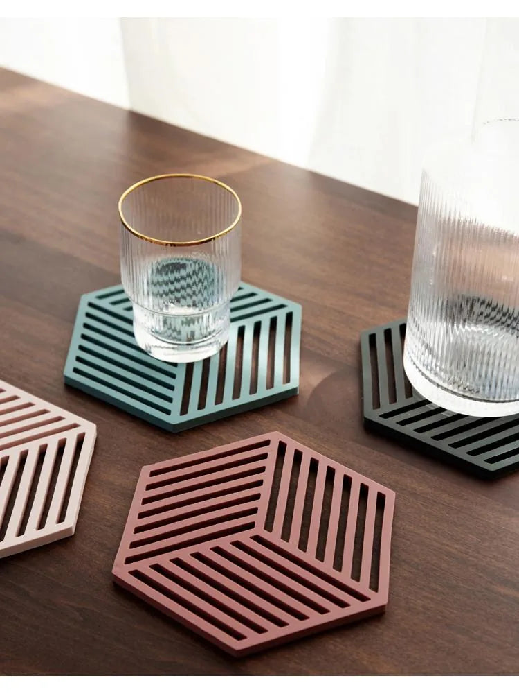 Deux sous-verres en silicone, sauge et terracotta, avec des verres posés dessus, et un troisième sous-verre rose au premier plan sur une table en bois.