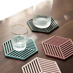 Deux sous-verres hexagonaux en silicone, vert sauge et rose poudré, avec des verres à eau côtelés posés dessus sur une table en bois foncé.