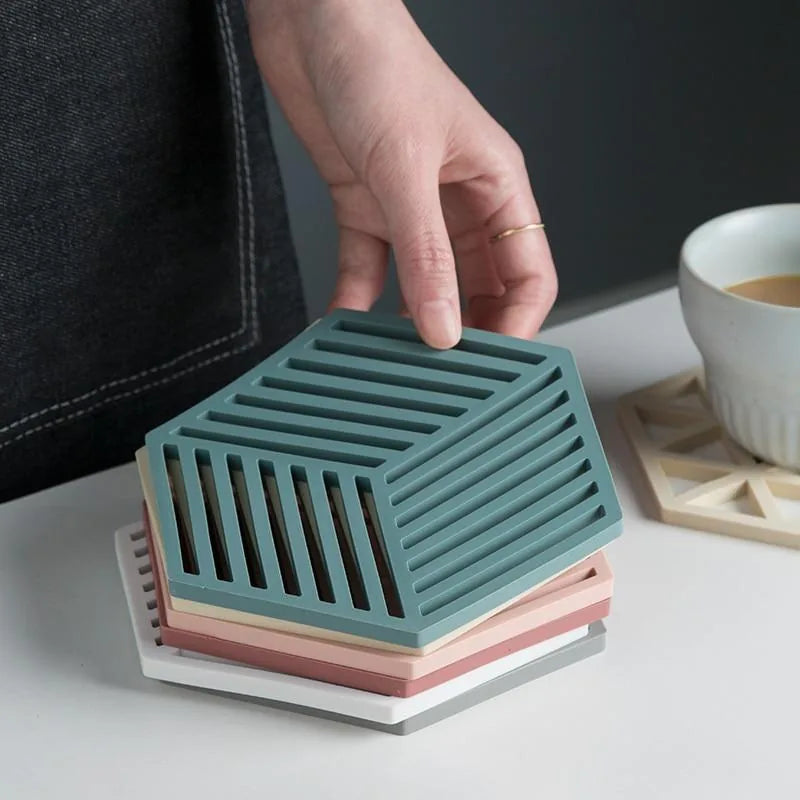 Une main soulève une pile de sous-verres hexagonaux en silicone de couleurs différentes empilés sur un plan de travail blanc.