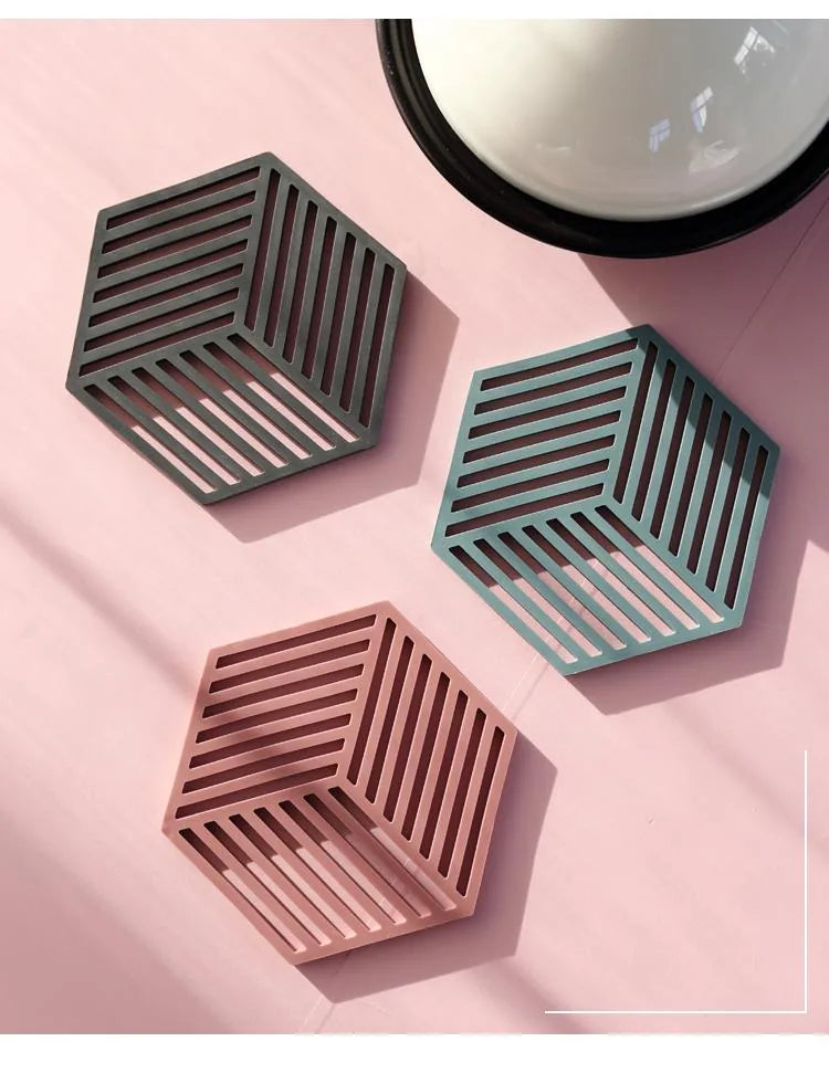 Vue de dessus de trois sous-verres hexagonaux en silicone vert foncé, sauge et rose à côté d'un bol noir sur un fond rose.