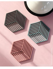 Vue de dessus de trois sous-verres hexagonaux en silicone vert foncé, sauge et rose à côté d'un bol noir sur un fond rose.
