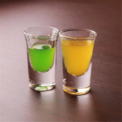 Gros plan sur deux verres à shot du lot, l'un rempli d'une liqueur verte et l'autre d'une liqueur orange, posés sur une table en bois foncé.