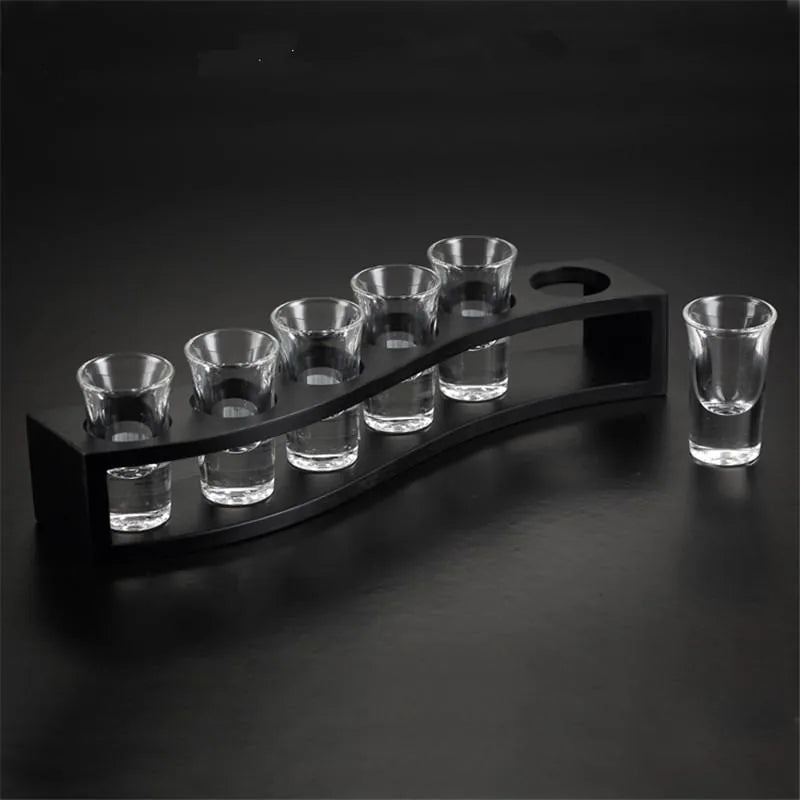 Support ondulé en bois noir avec six verres à shot vides et un septième verre posé séparément, sur fond noir dégradé.