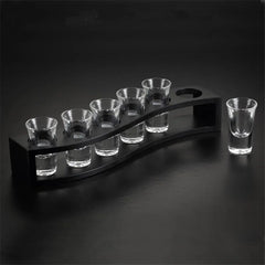 Support ondulé en bois noir avec six verres à shot vides et un septième verre posé séparément, sur fond noir dégradé.