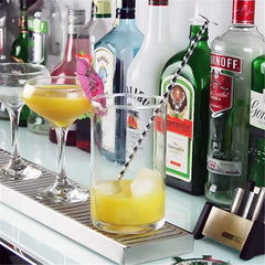 Le verre à mélange rempli de glaçons et de cocktail orangé posé sur un comptoir de bar entouré de bouteilles de spiritueux et d'une coupe garnie d'une fleur.