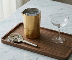 Verre à mélange doré accompagné d'une passoire à glaçons et d'une coupe à cocktail, posés sur un plateau en bois sur un plan de marbre.