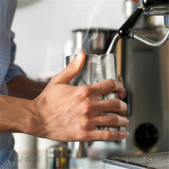 Main tenant le verre à mélange en inox sous la buse vapeur d'une machine à café, avec de la vapeur visible