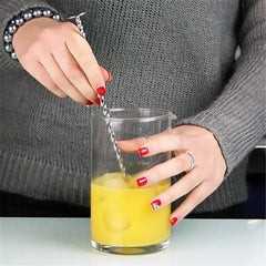 Gros plan sur des mains aux ongles vernis rouge remuant un cocktail à l'orange dans un verre à mélange à l'aide d'une cuillère de bar torsadée.