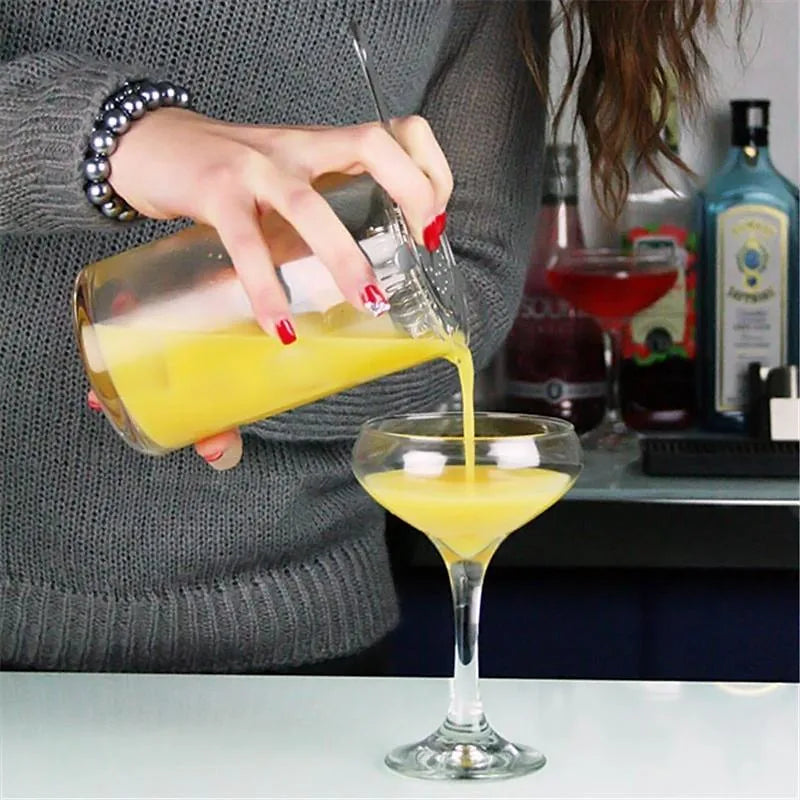 Une barmaid verse un cocktail orangé depuis le verre à mélange dans une coupe à cocktail, sur un comptoir de bar.
