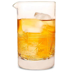 Verre à mélange vintage en cristal taillé rempli de whisky ambré et d'un gros glaçon, photographié sur fond blanc.