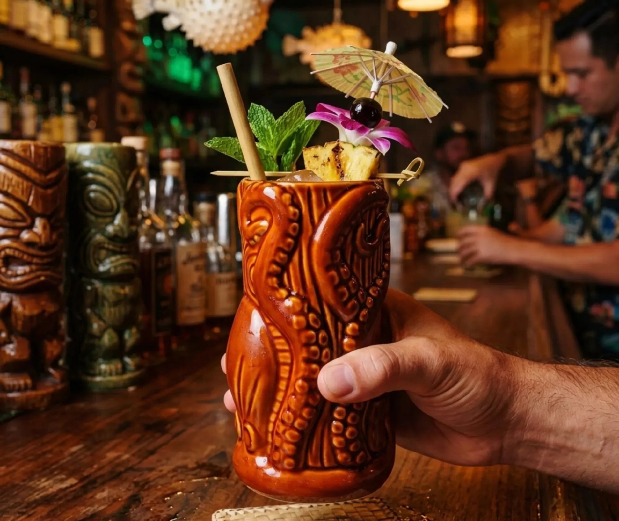 Verre à rhum Tiki tenu à la main au comptoir d'un bar, garni d'un parasol en papier, d'une cerise et de menthe fraîche