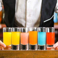 Série de six verres à shot longs remplis de cocktails multicolores tenus par un barman