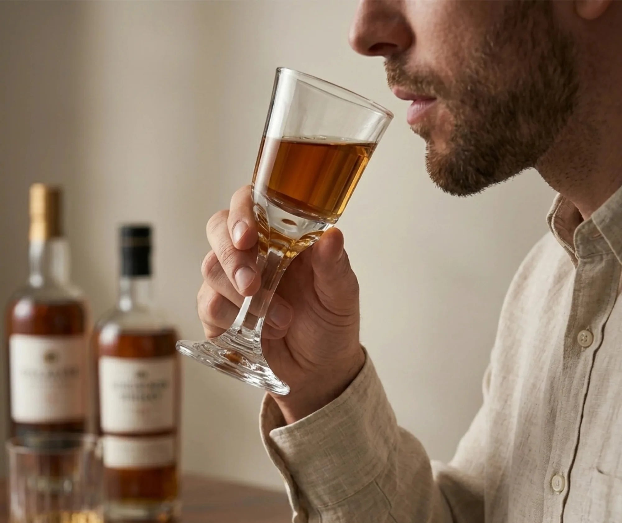 Un homme dégustant un rhum ambré dans un verre à shot à pied, bouteilles de spiritueux floues en arrière-plan.