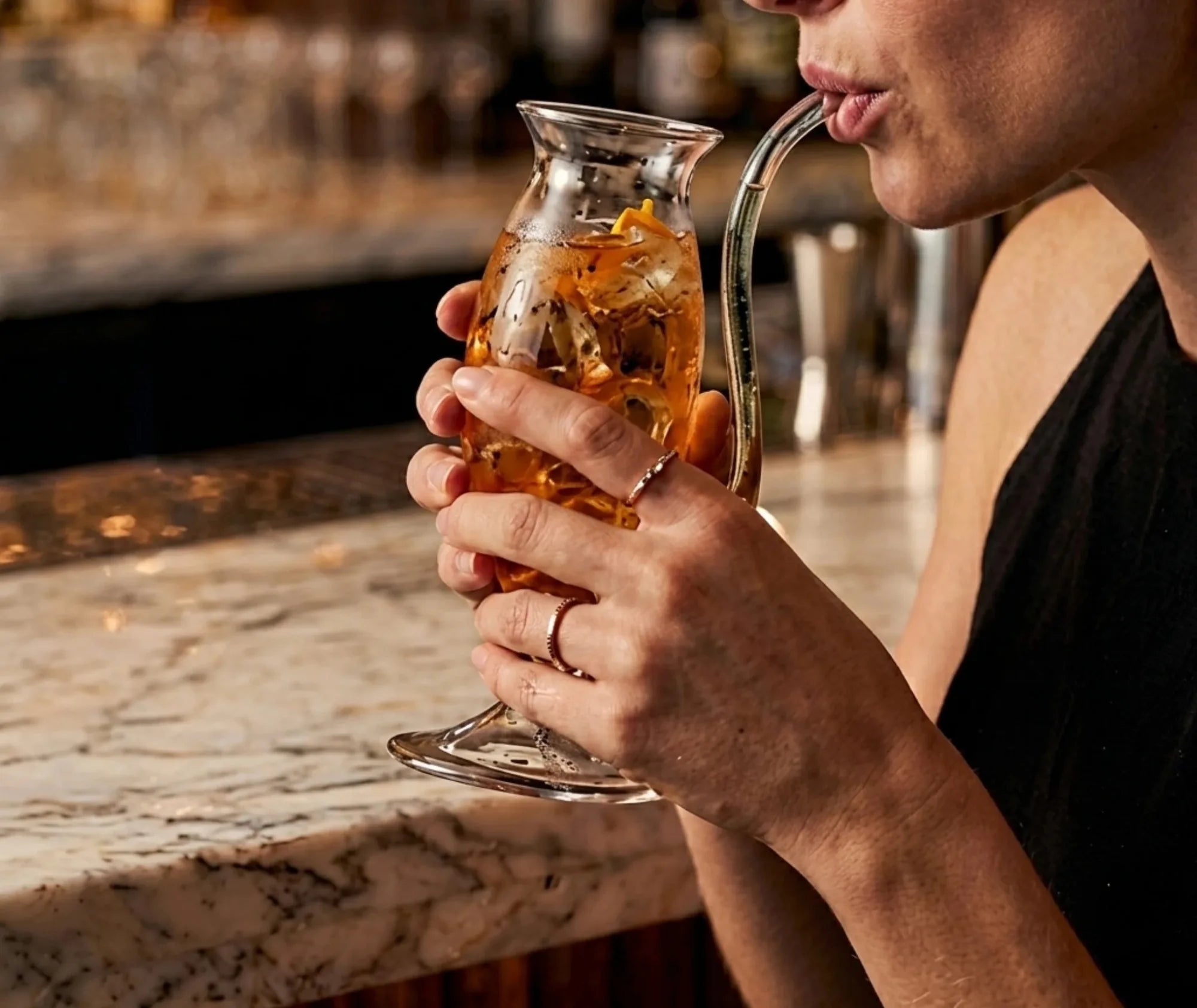 Femme dégustant un cocktail ambré directement au bec verseur du verre à trompe, accoudée à un bar en marbre.