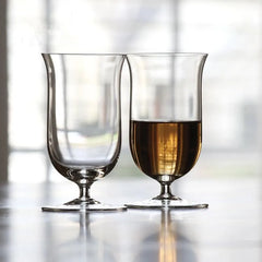 Deux verres à whisky à pied côte à côte sur un comptoir clair, l'un rempli de whisky, avec une fenêtre lumineuse en arrière-plan.