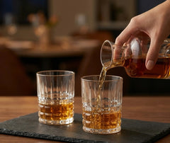 Whisky versé depuis une carafe dans deux verres à whisky de bar posés sur un plateau en ardoise.