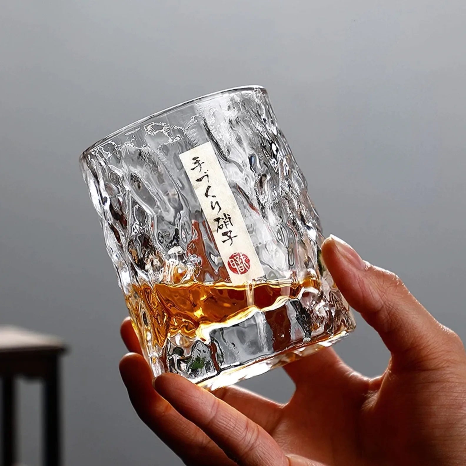 Gros plan sur un verre à whisky japonais tenu en main, rempli de whisky ambré, mettant en valeur la texture martelée du verre et son étiquette.