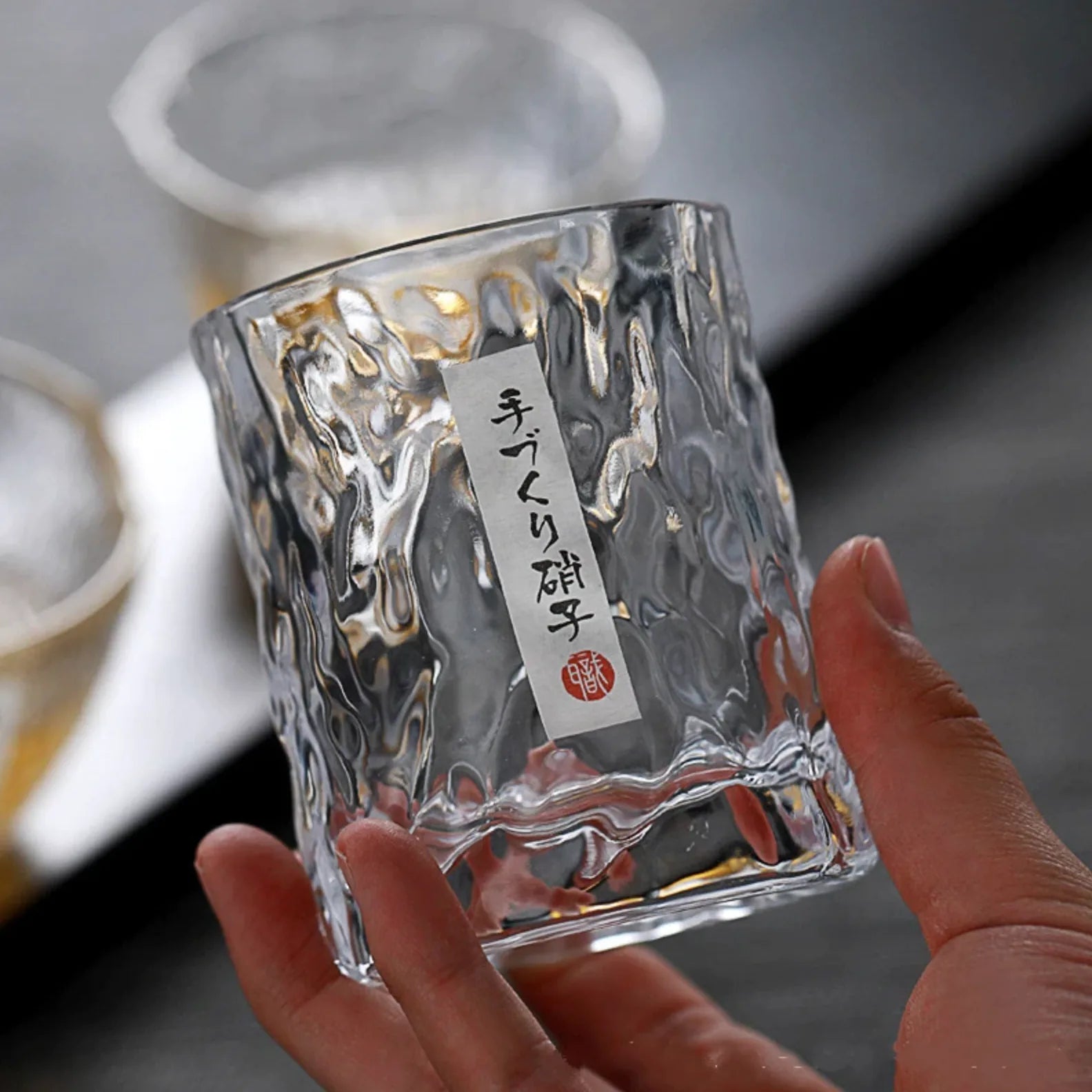 Gros plan sur la texture martelée du verre à whisky japonais tenu en main, avec son étiquette artisanale japonaise portant la mention verre fait main.