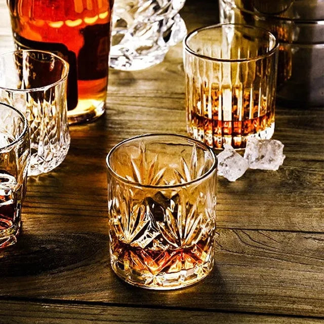 Plusieurs verres à whisky vintage aux motifs variés disposés sur une table en bois avec bouteille et glaçons.