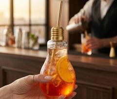 Verre ampoule à cocktail contenant un apéritif orange avec glaçons et une tranche d'orange, tenu à la main dans une ambiance de bar.