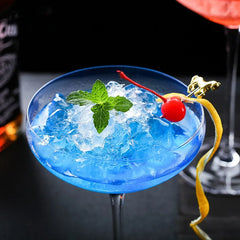 Cocktail bleu servi dans un verre à cocktail coupe avec glace pilée, menthe fraîche et cerise confite, dans une ambiance de bar sombre.