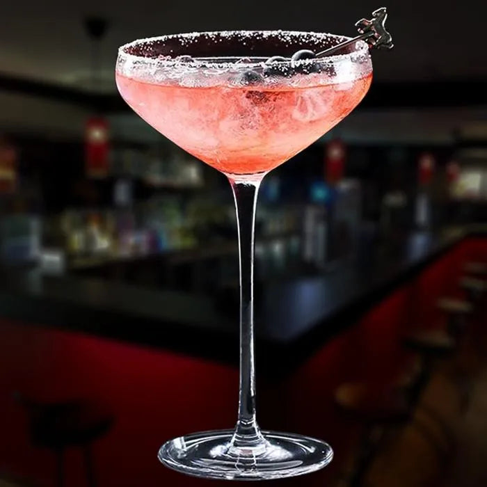 Cocktail rose servi dans un verre à cocktail coupe, posé sur le comptoir d'un bar sombre en arrière-plan.