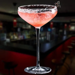 Cocktail rose servi dans un verre à cocktail coupe, posé sur le comptoir d'un bar sombre en arrière-plan.