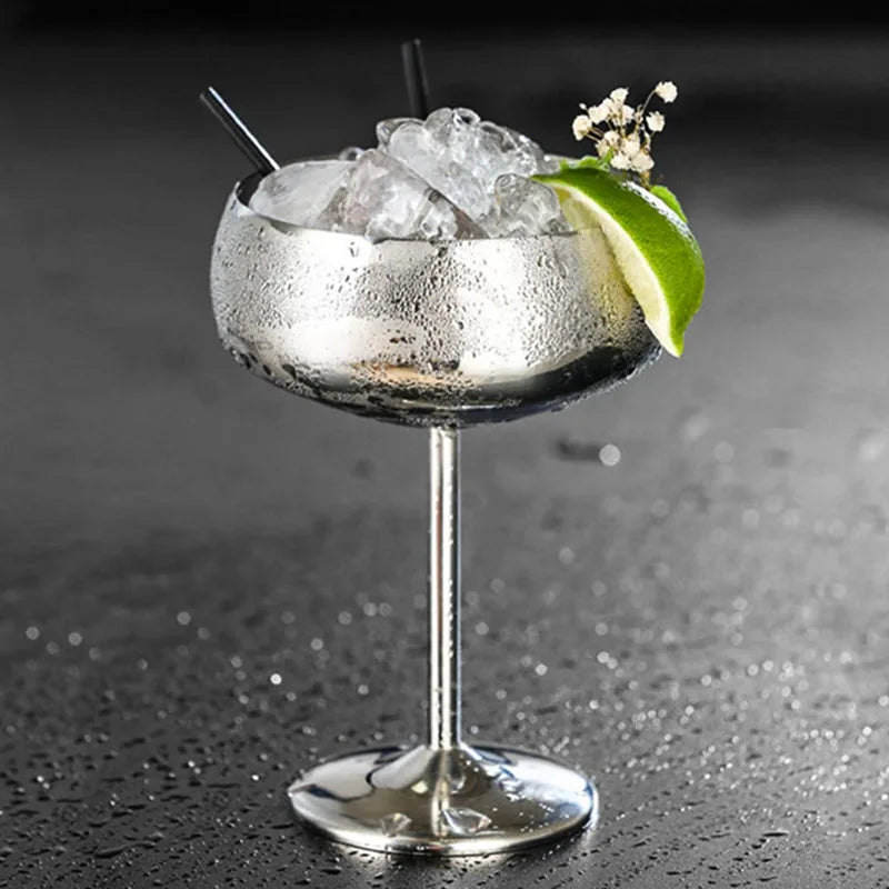 Verre à cocktail coupe argenté avec deux pailles noires, glaçons, quartier de citron vert et fleurs séchées, sur fond gris.