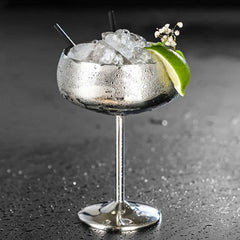 Verre à cocktail coupe argenté avec deux pailles noires, glaçons, quartier de citron vert et fleurs séchées, sur fond gris.