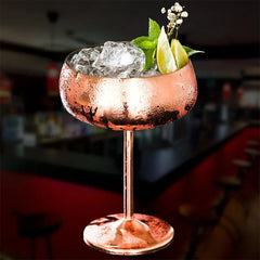 Verre à cocktail coupe en cuivre rempli de glaçons, garni de menthe fraîche, de fleurs et de quartiers de citron vert, sur fond sombre de bar.