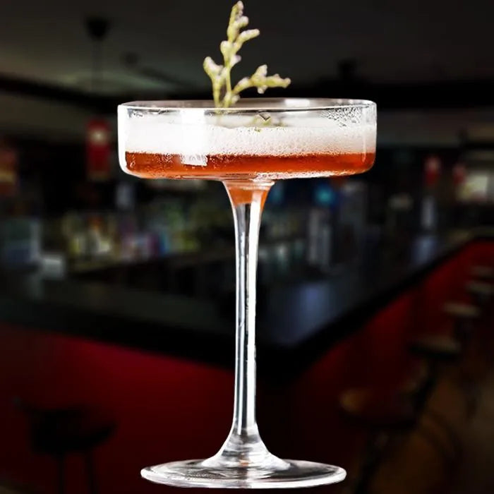 Verre à cocktail coupe plate rempli d'un cocktail rouge ambré mousseux, garni d'une branche d'herbe aromatique, dans une ambiance de bar en arrière-plan flou.