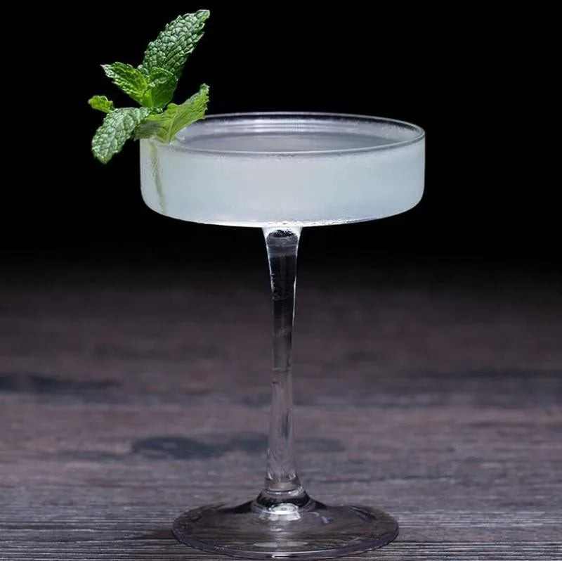 Gros plan d'un verre à cocktail coupe plate rempli d'un cocktail blanc givré, décoré d'un brin de menthe fraîche, sur fond de bois sombre.