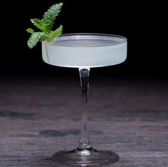 Gros plan d'un verre à cocktail coupe plate rempli d'un cocktail blanc givré, décoré d'un brin de menthe fraîche, sur fond de bois sombre.
