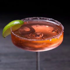 Verre à cocktail coupe plate au bord givré de sel rosé, rempli d'un cocktail orangé avec des glaçons et garni d'un quartier de citron vert.