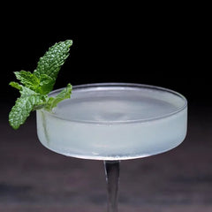 Gros plan d'un verre à cocktail coupe plate rempli d'un cocktail transparent légèrement givré, orné d'un brin de menthe, sur fond sombre.