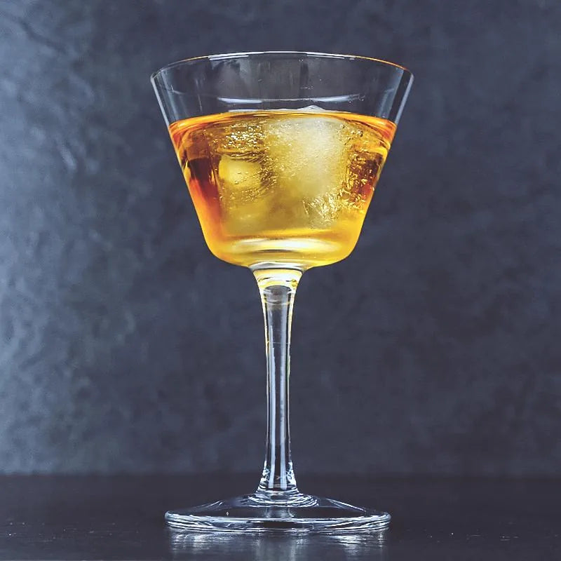 Verre à cocktail coupe vintage rempli d'un cocktail ambré avec un glaçon, photographié en studio sur fond gris-bleu texturé.