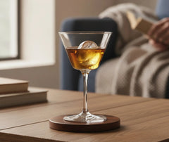 Verre à cocktail coupe vintage contenant un cocktail ambré avec un glaçon sphérique, posé sur une table basse en bois dans un salon cosy.