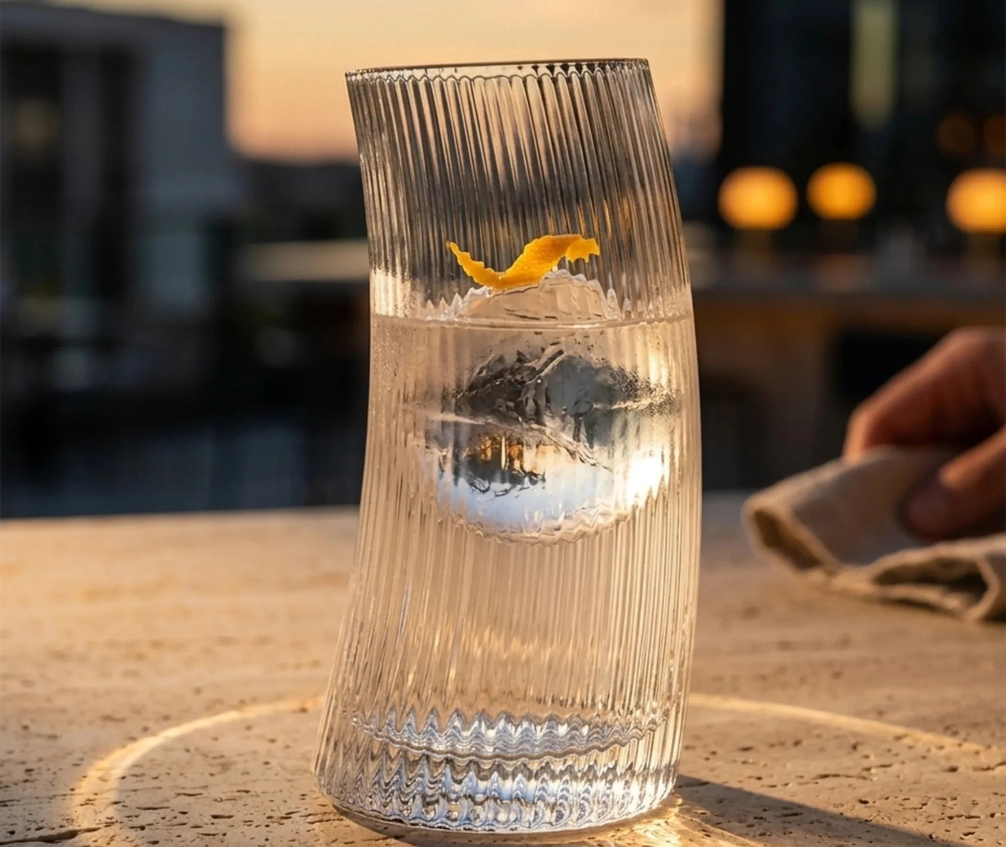 Verre à cocktail courbé posé sur une table en terrasse au coucher de soleil, garni d'un zeste d'orange