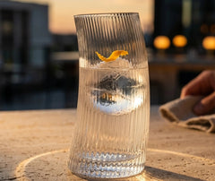 Verre à cocktail courbé posé sur une table en terrasse au coucher de soleil, garni d'un zeste d'orange