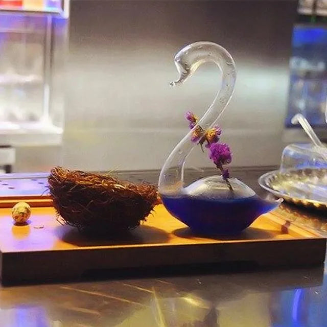 Verre à cocktail cygne rempli d'un cocktail violet avec fleurs séchées, posé sur un plateau en bois à côté d'un nid décoratif