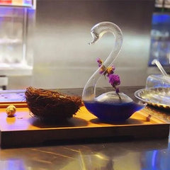 Verre à cocktail cygne rempli d'un cocktail violet avec fleurs séchées, posé sur un plateau en bois à côté d'un nid décoratif