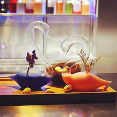 Duo de verres à cocktail cygne, l'un rempli d'un cocktail violet et l'autre d'un cocktail orange, présentés sur un plateau en bois avec un nid décoratif et des œufs de caille