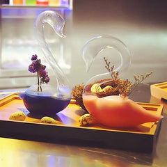 Duo de verres à cocktail cygne, bleu-violet et orange, présentés côte à côte sur un plateau en bois avec nid décoratif et œufs de caille, vue large