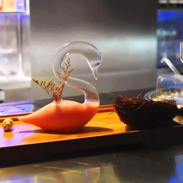 Verre à cocktail cygne rempli d'un cocktail orange, posé sur un plateau en bois à côté d'un nid décoratif
