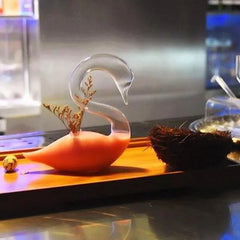 Verre à cocktail cygne rempli d'un cocktail orange, posé sur un plateau en bois à côté d'un nid décoratif