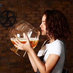 Femme aux cheveux bouclés vue de profil tenant un verre à cocktail géant rempli de bière blonde, devant un mur de briques