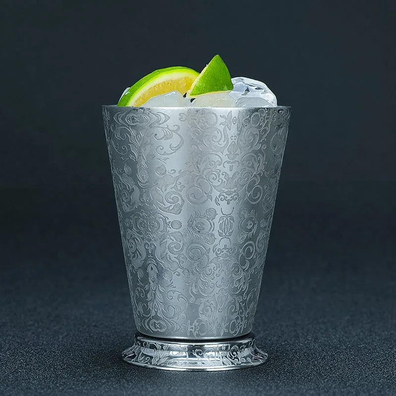 Verre à cocktail Gourmandise finition Cachemire au motif gravé, rempli de glace avec des rondelles de citron vert et jaune, sur fond noir.