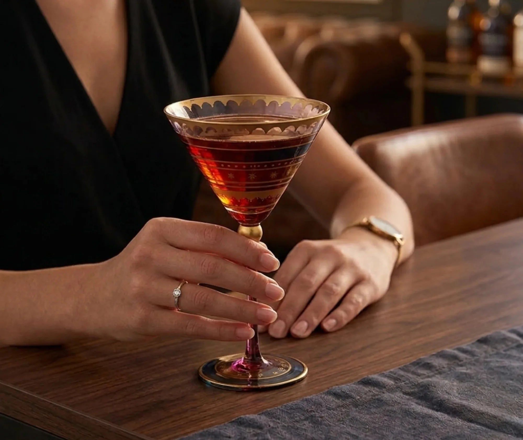 Femme tenant un verre martini à dorures rempli d'un cocktail rouge, accoudée à un bar en bois