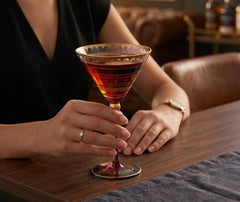 Femme tenant un verre martini à dorures rempli d'un cocktail rouge, accoudée à un bar en bois