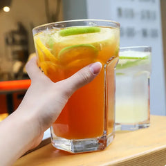 Verre à cocktail Mojito XL tenu en main rempli de thé glacé ambré aux rondelles de citron vert, sur une table en bois clair